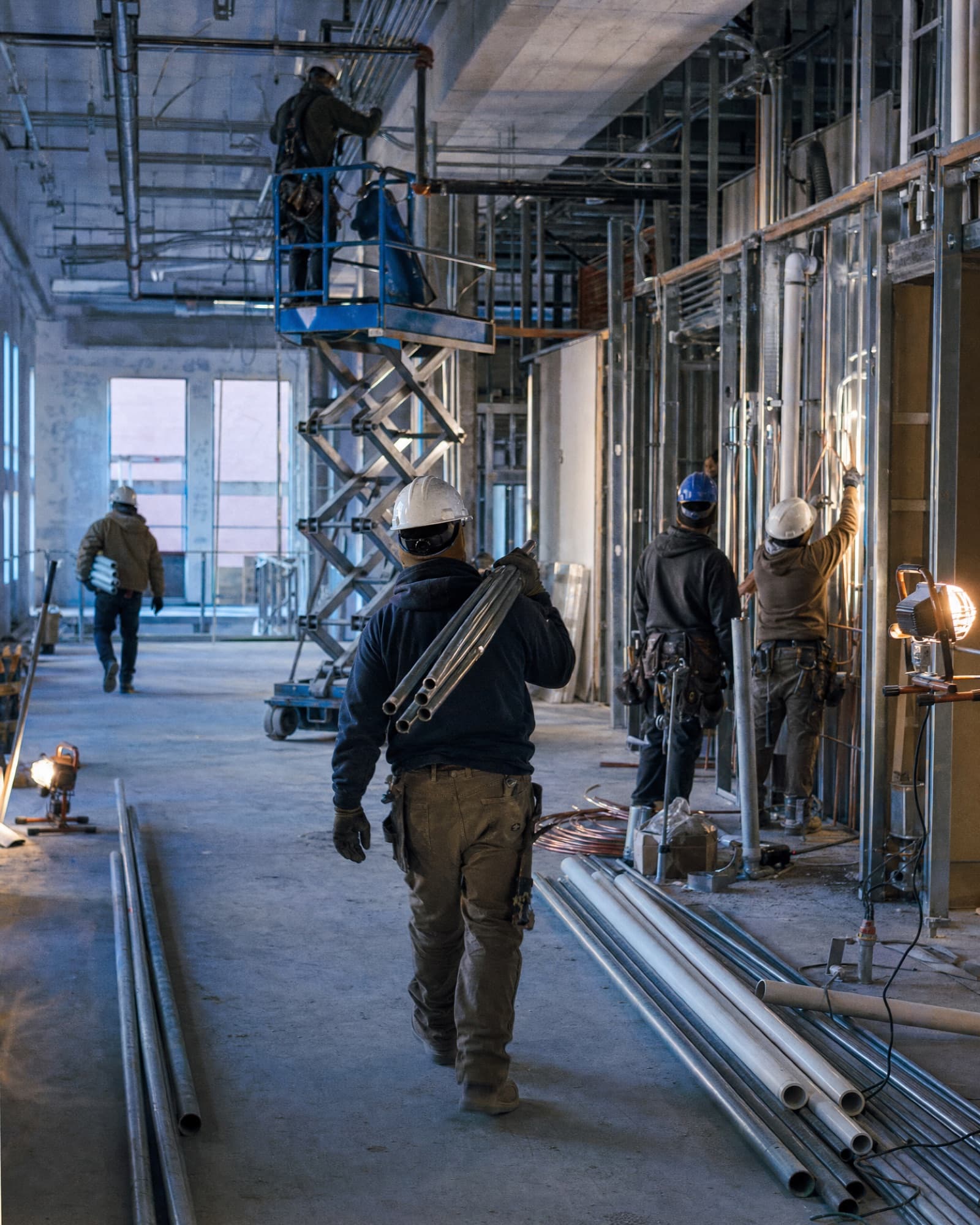 A trades crew roughing in a commercial building: conduit and pipe through open studs, a man on a scissor lift overhead